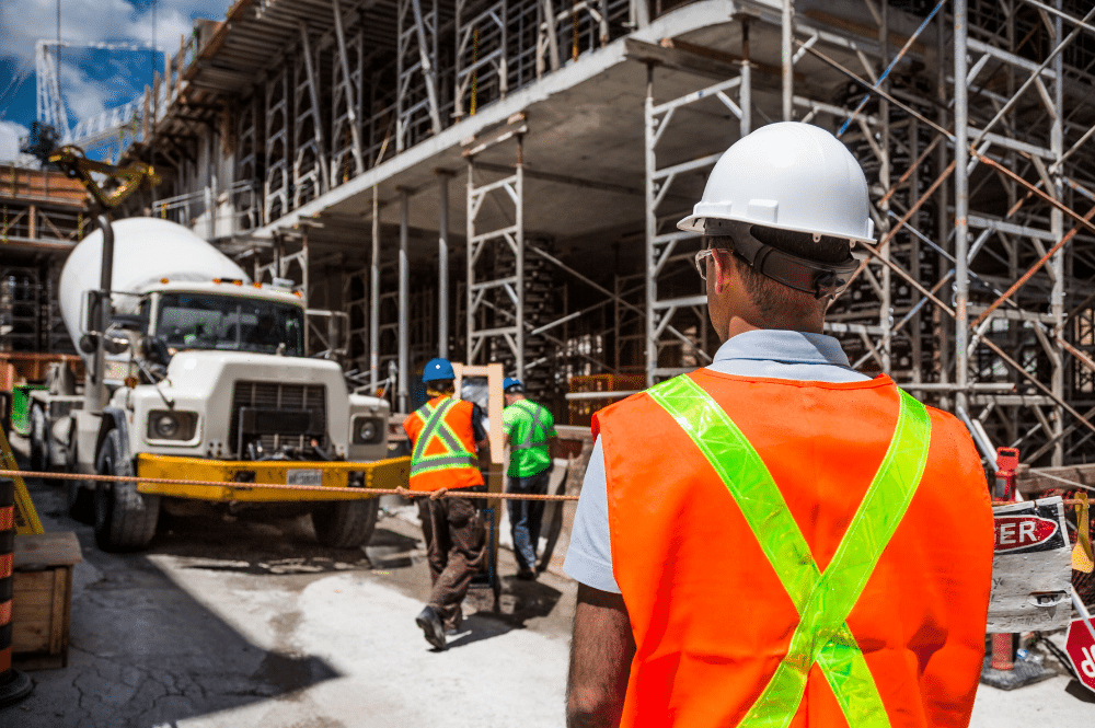 A construction worker in safety gear stands confidently in front of a bustling construction site, ensuring safety regulations are met.